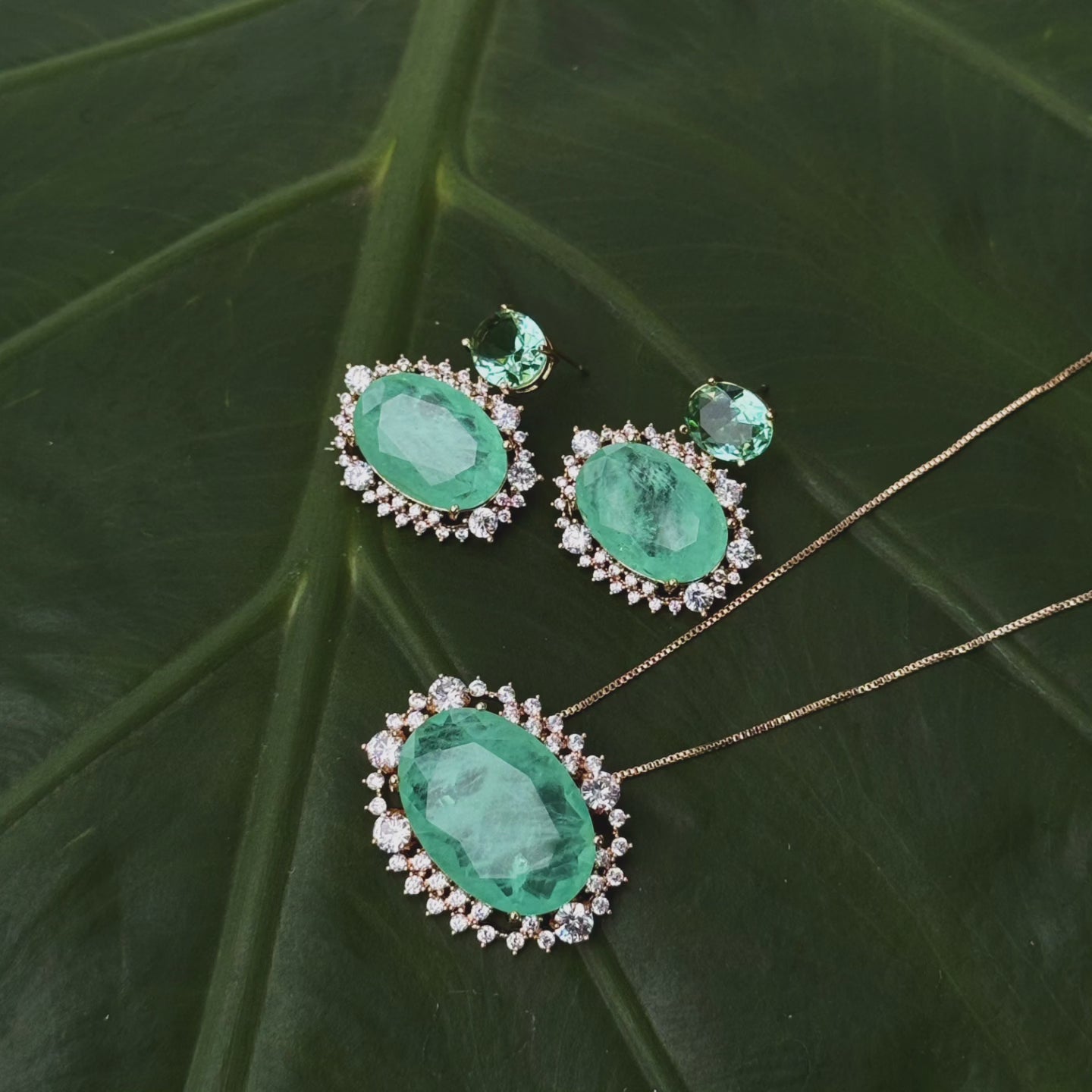 A luxurious jewelry set displayed on a green leaf background, featuring an 18k gold-plated necklace with a vibrant fusion tourmaline stone surrounded by sparkling accents, paired with matching earrings.