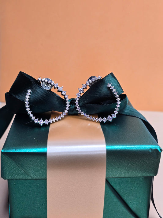 A pair of earrings shaped like irregular circle, embellished with different shaped Zircons all around the earring, displayed on a green gift box with a white ribbon tied in a bow. The background is out-of-focus with peach or light orange tones, highlighting the playful and elegant design of the earrings