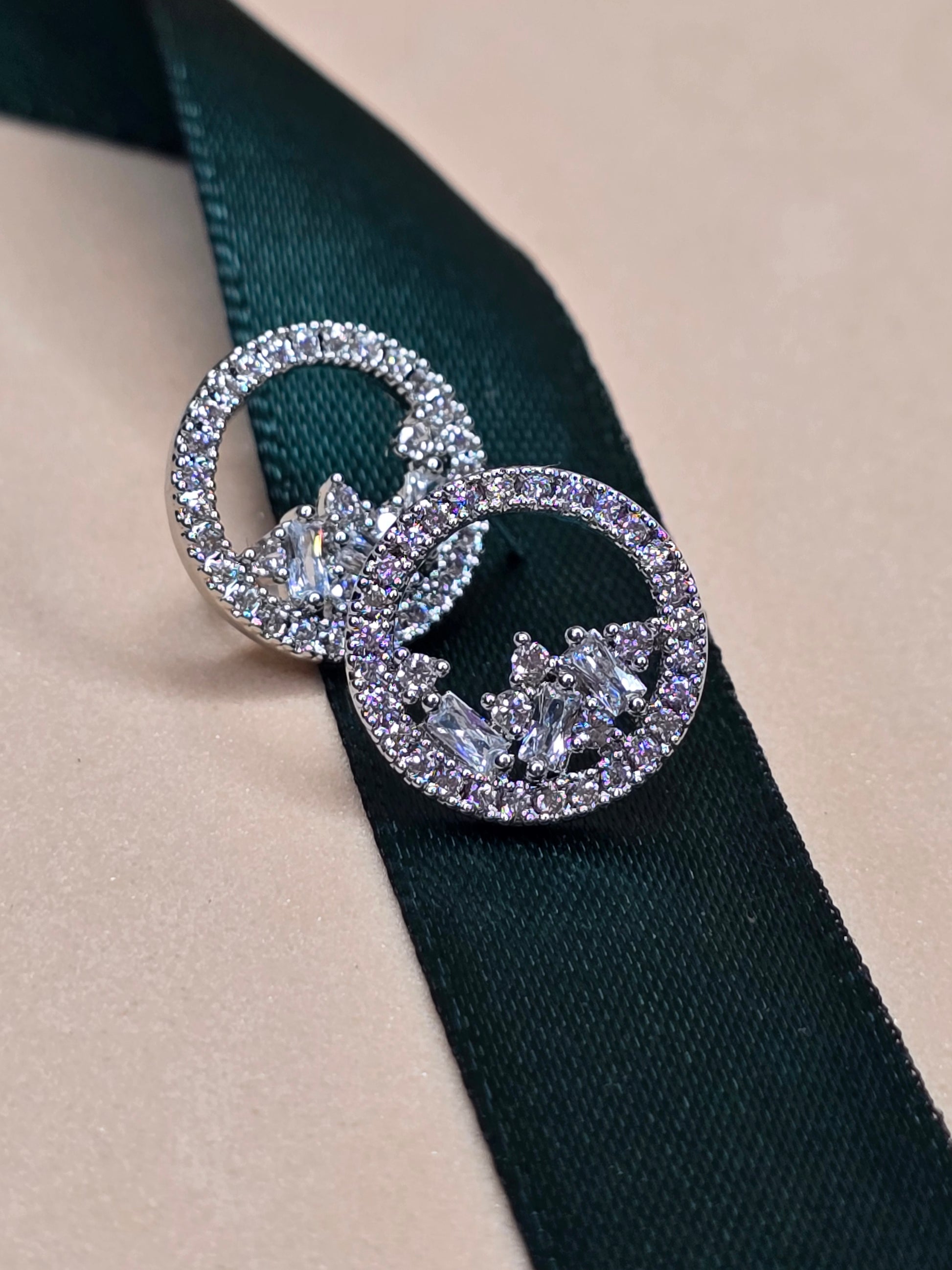 A pair of circular rhodium-plated earrings with embedded zircon gemstones shaped as hearts, resting on a dark green ribbon against a light beige background. The earrings feature smaller circles inside the larger ones, both lined with sparkling gemstones and intricate silver-colored metalwork at the center.