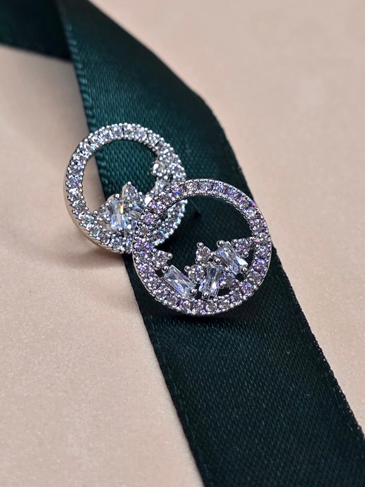 A pair of circular rhodium-plated earrings with embedded zircon gemstones shaped as hearts, resting on a dark green ribbon against a light beige background. The earrings feature smaller circles inside the larger ones, both lined with sparkling gemstones and intricate silver-colored metalwork at the center.