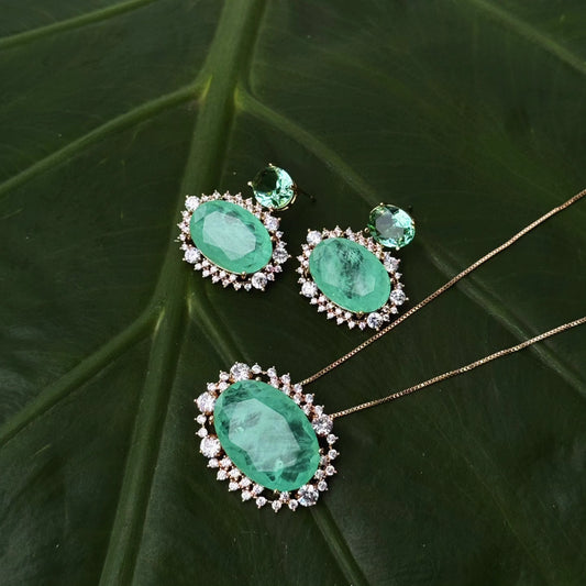 A luxurious jewelry set displayed on a green leaf background, featuring an 18k gold-plated necklace with a vibrant fusion tourmaline stone surrounded by sparkling accents, paired with matching earrings.