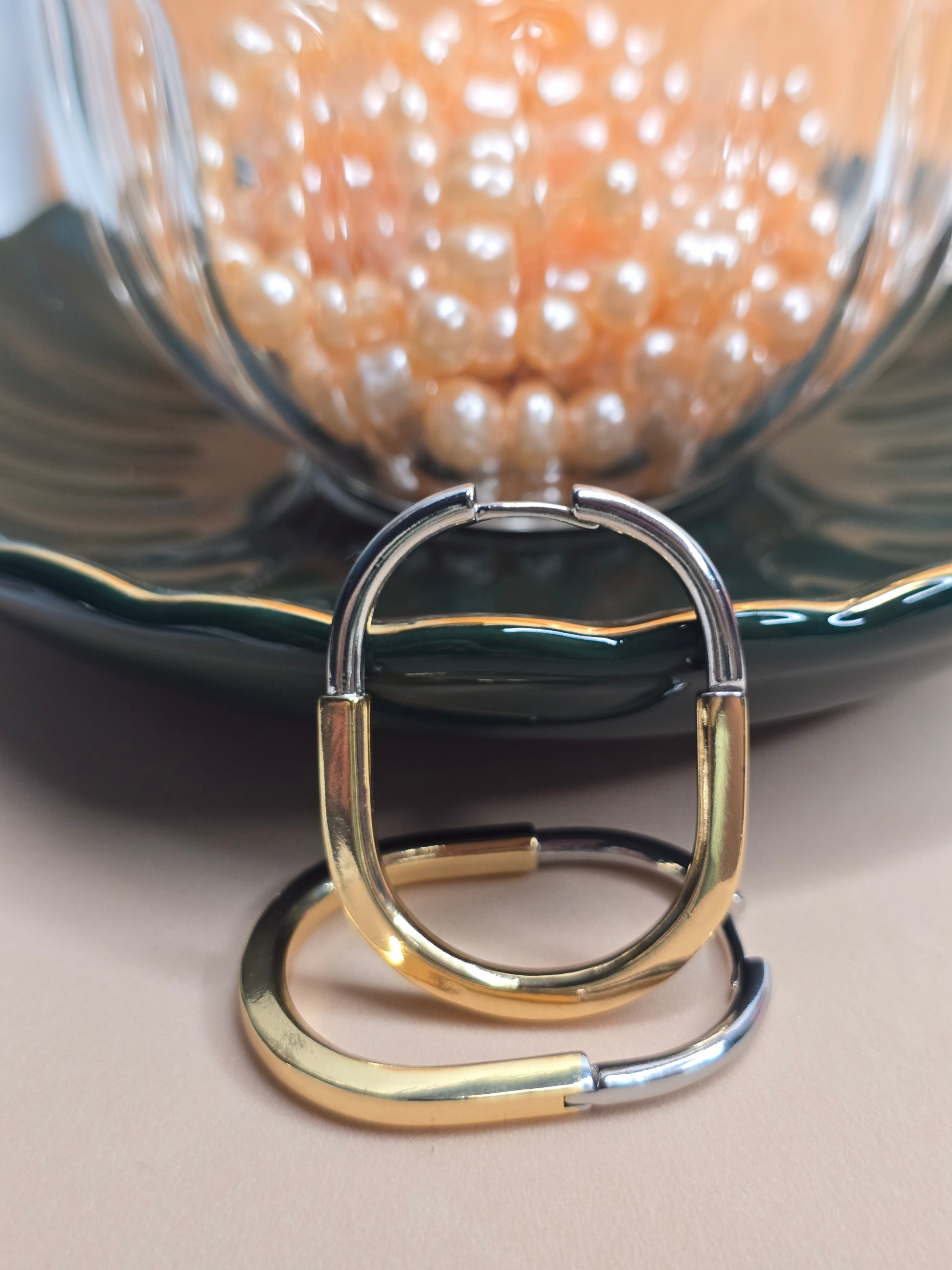 A pair of oval hoop earrings with a dual-tone design, half gold and half silver-colored, resting on a green palte with a glass tea cup with pearls in them.