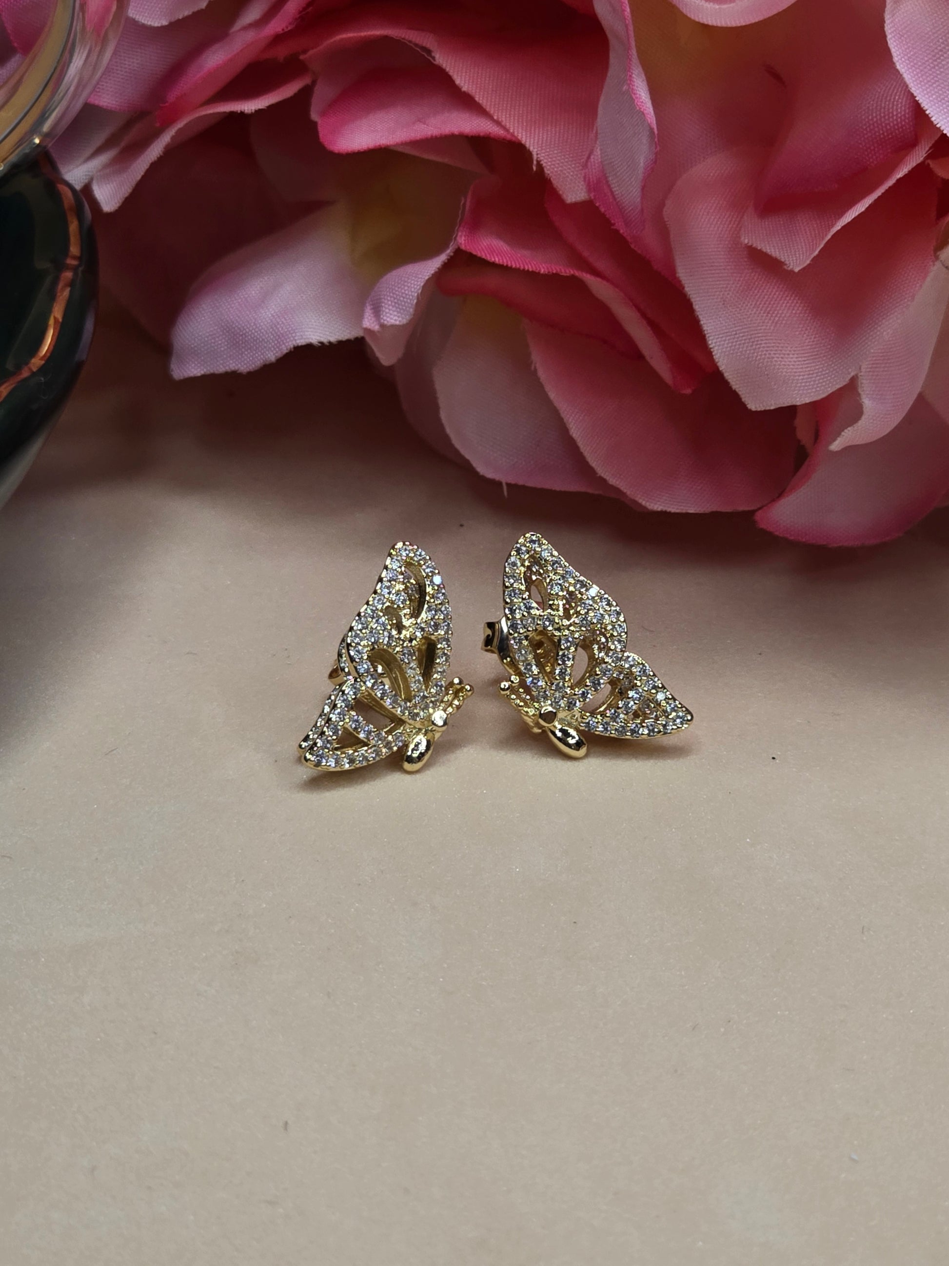 A pair of butterfly-shaped earrings with a gold-tone finish and embellished with numerous small, clear zircons, placed on a light peach surface with a blurred floral element in the background.