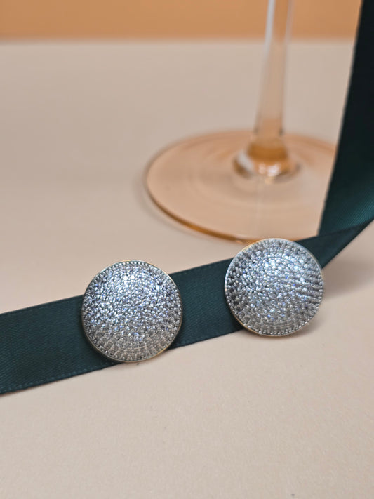 A pair of round, 18k gold-plated earrings with numerous small zircons embedded on their surface, displayed against a soft beige background and resting on a dark green ribbon