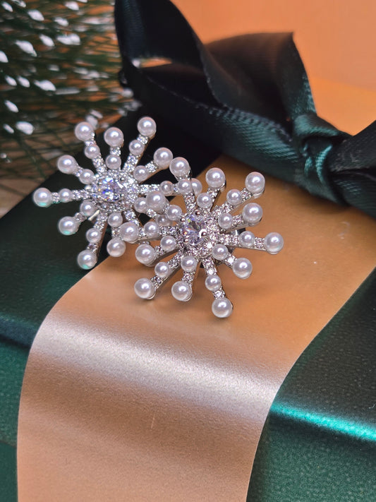 A pair of snowflake-shaped earrings with small pearls and crystals on their arms, resting on a green and bronze satin ribbon.