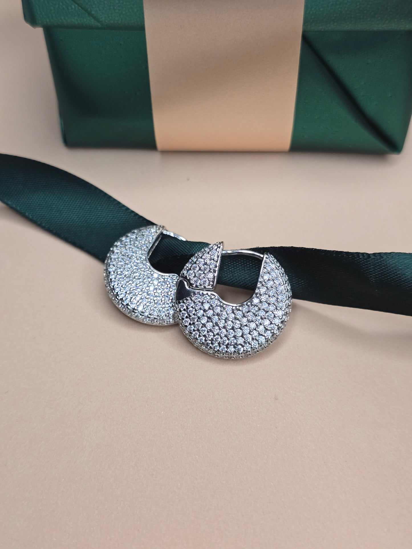 A pair of crescent-shaped hoop earrings encrusted with numerous tiny crystals are displayed in front of a green gift box with a dark green ribbon on a beige surface. The earrings sparkle against the neutral tones, suggesting sophistication and luxury