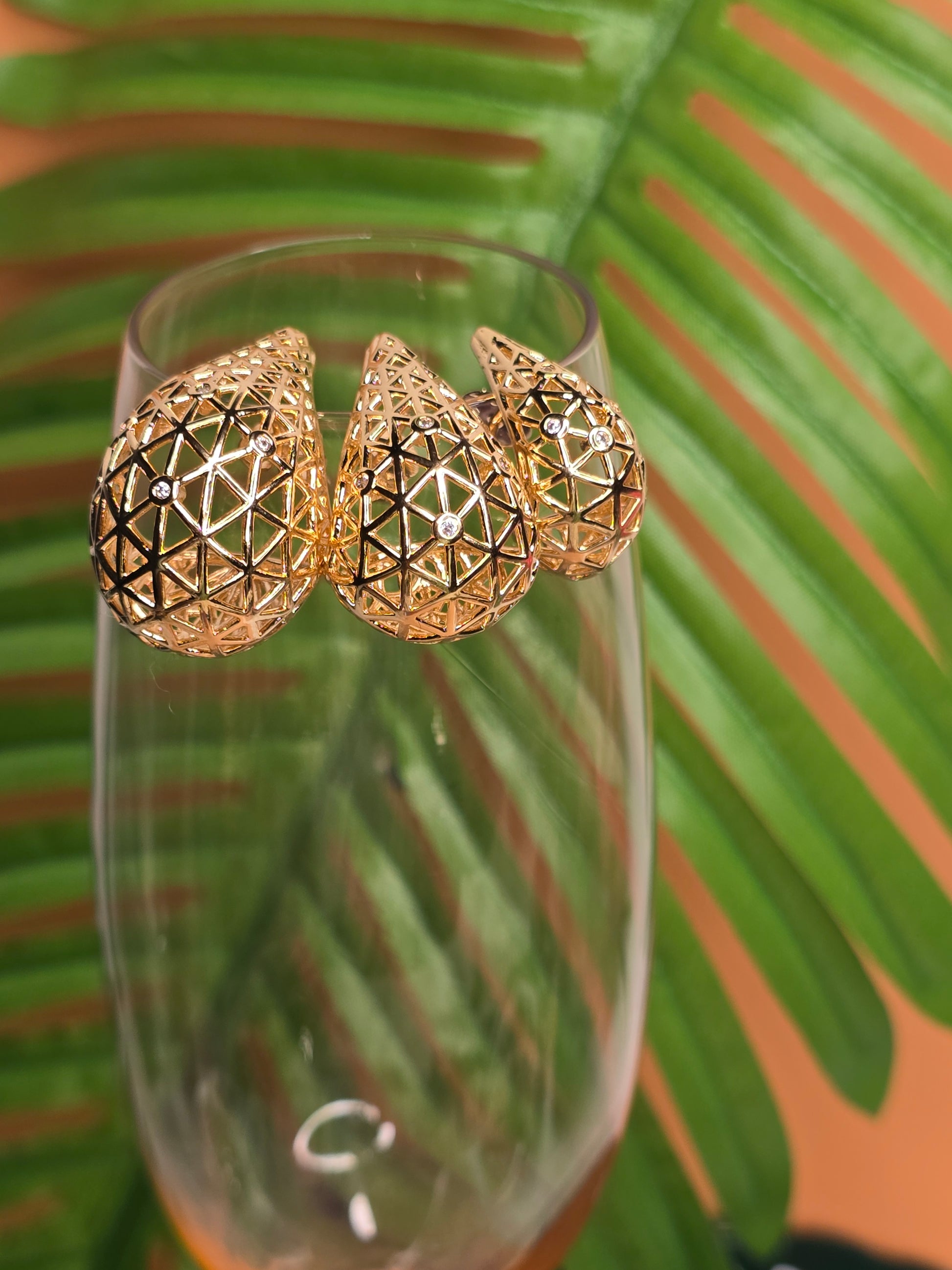 A close-up view of 3 sizes of the gold geometric patterned teardrop earrings leaning on the edge of a peach faceted glass cup with soft-focus green plant in the background