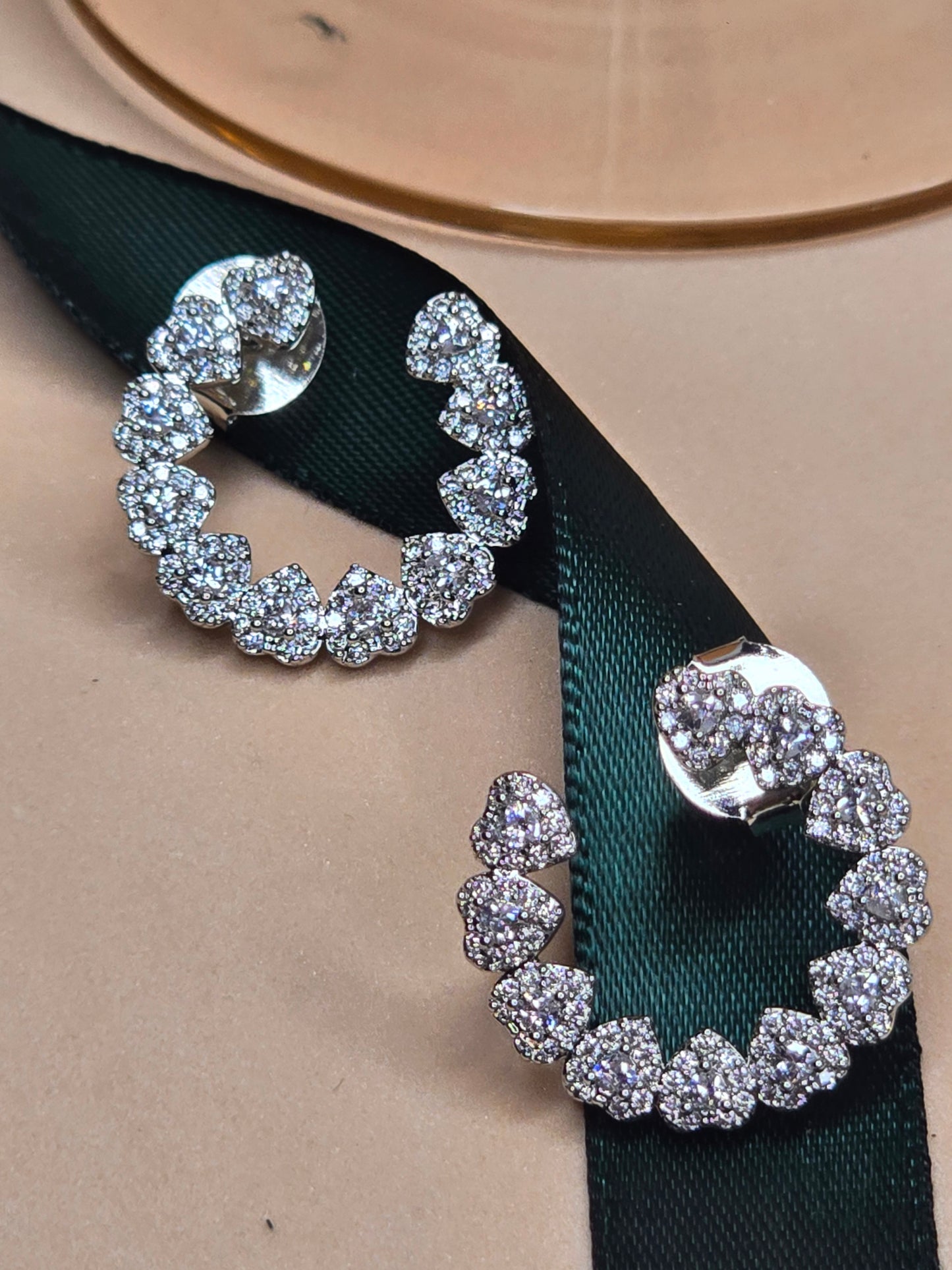 A pair of rhodium-plated hoop earrings encrusted with sparkling zircon stones, displayed on a soft, peach-toned surface. The earrings shine brightly, suggesting elegance and luxury.