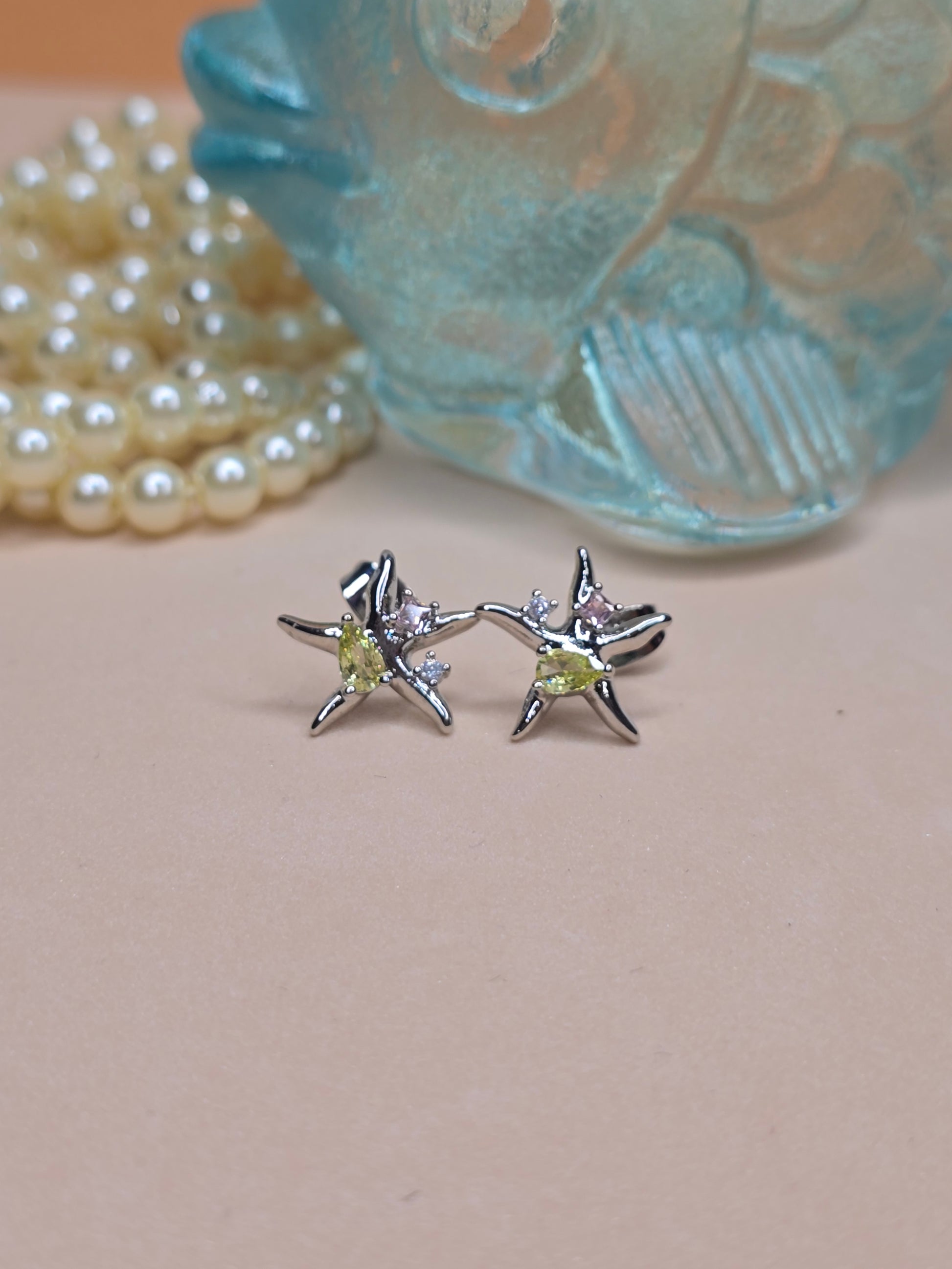 A pair of star-shaped earrings with a central yellow stone and smaller clear stones extending along the star’s arms, placed on a light pink surface with a blurred decorative glass item and a string of pearls in the background.