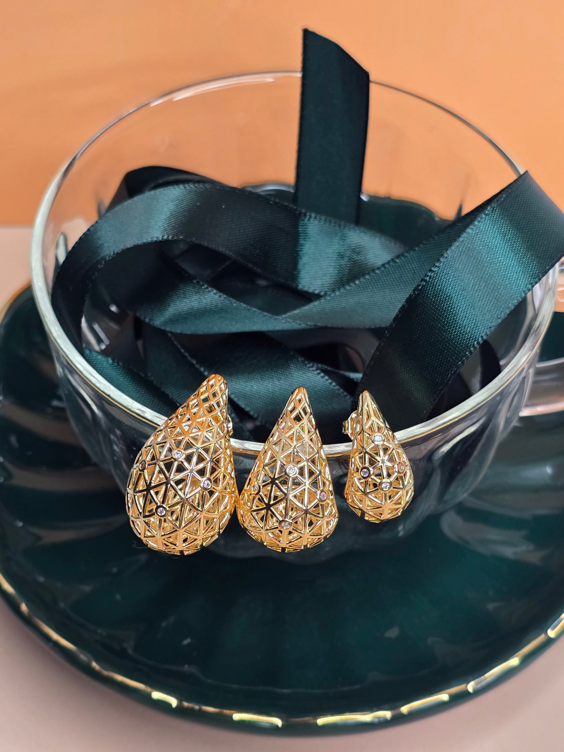 A close-up view of gold geometric patterned teardrop earrings leaning on the edge of a clear faceted glass cup, with green ribbon inside of it with soft-focus peach background