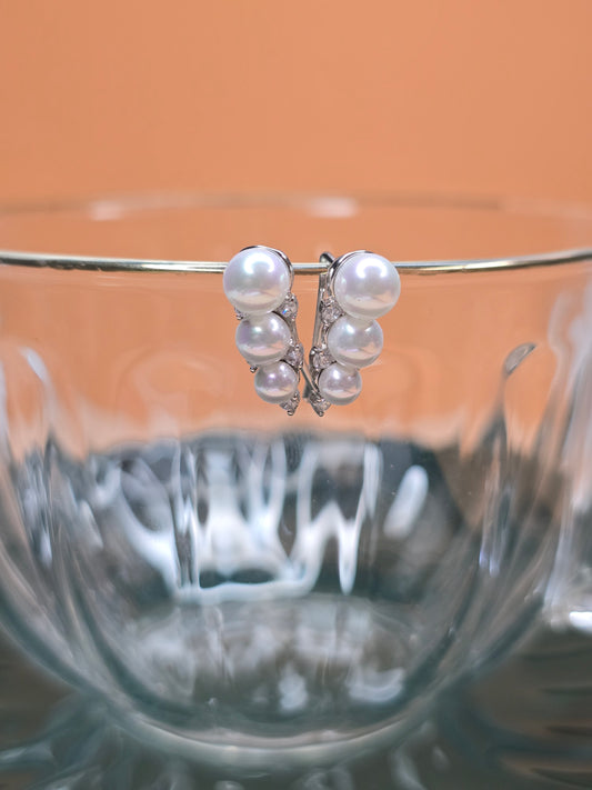 A pair of pearl earrings with a silver setting in cuff style, delicately perched on the rim of a transparent glass. The background is a soft, peach-toned hue that contrasts with the clear and reflective surface of the glass, highlighting the lustrous quality.