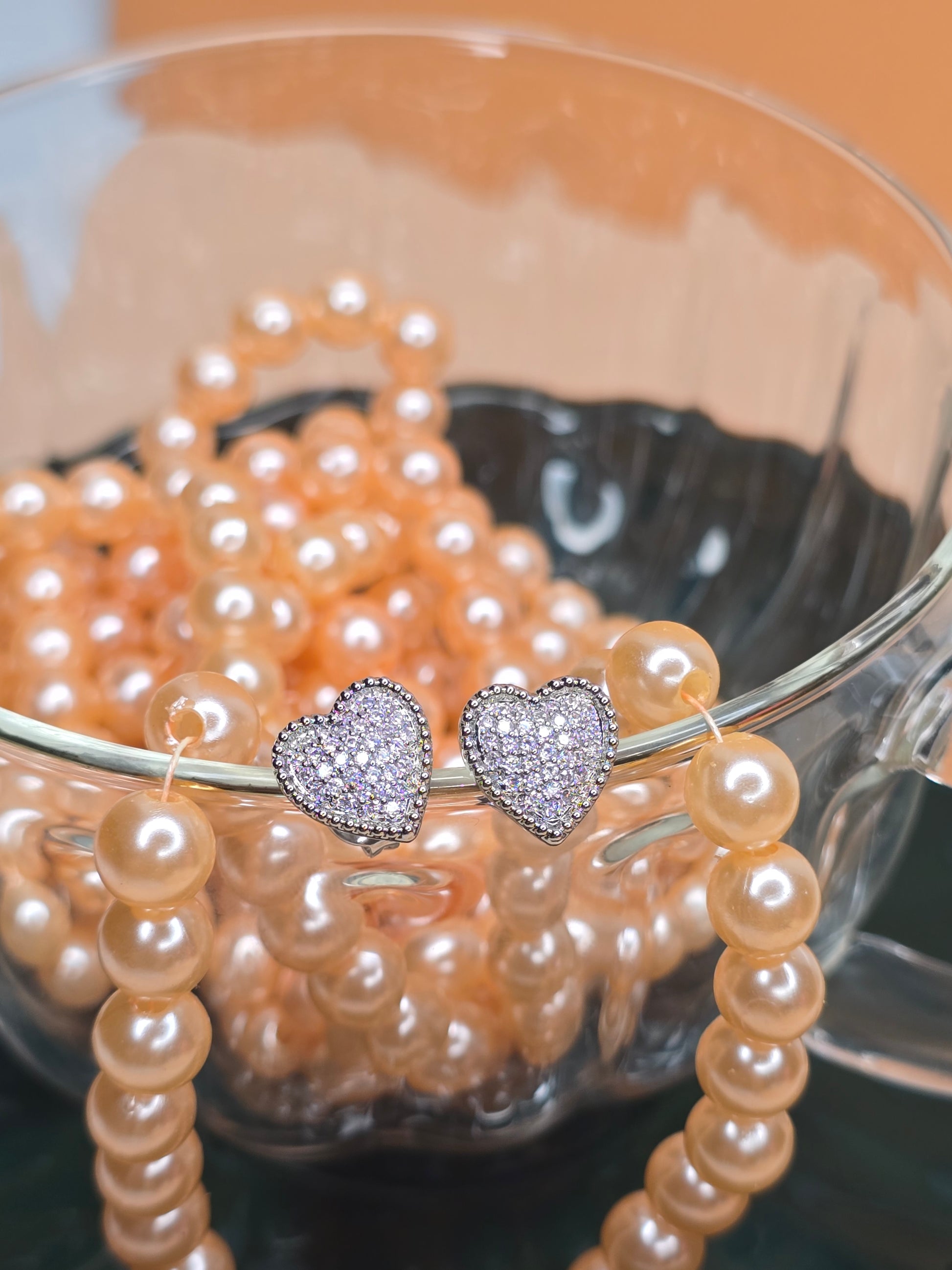 A pair of heart-shaped earrings with diamond embellishments placed on top of a string of pearls inside a clear glass bowl. The sparkling earrings contrast beautifully with the soft luster of the pearls, suggesting elegance and luxury